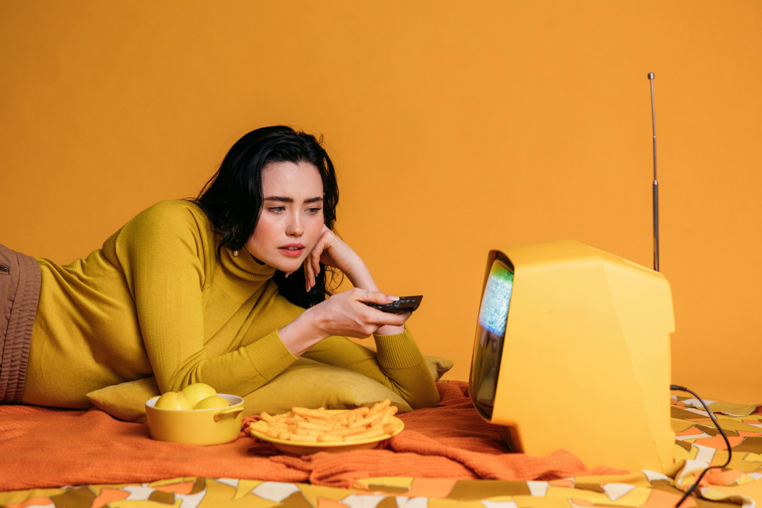 A influencer lady wearing over neck t-shirt watching a vintage arial television laying on mat. Entire image is in yellow theme.